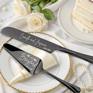 Könnte beinhalten: Schwarzes Hochzeitstortenmesser- und -server-Set mit gravierten Namen und Hochzeitsdatum. "Camille und Jasmine" und "Mr. und Mrs. Brayford" sind in die Messer graviert. Die Messer liegen auf einem weißen Teller mit einer weißen Serviette und Perlen.