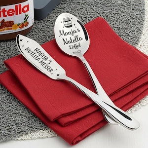 May include: A stainless steel spoon and spreader set with engraved text. The spoon reads "Monja's Nutella Löffel" and the spreader reads "Magija's Nutella Messer." Both are on a red napkin, with a Nutella jar visible in the background.
