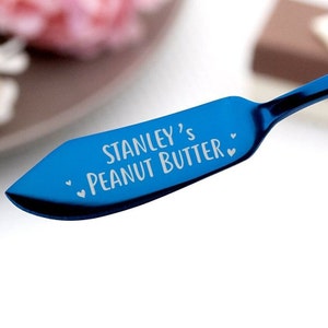 May include: A blue stainless steel butter knife with the text "Stanley's Peanut Butter" engraved on the blade. Two small hearts are engraved on either side of the text.