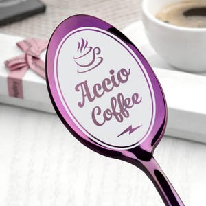 May include: A purple coffee spoon with the words "Accio Coffee" and a coffee cup graphic. The spoon has a shiny finish. The background includes a coffee cup and a gift box, both out of focus.