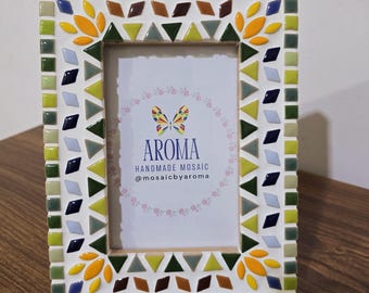 May include: A white picture frame with a mosaic design. The frame is decorated with yellow, green, blue, and brown tiles. The frame has a small rectangular opening for a picture. The text "AROMA HANDMADE MOSAIC @mosaicbyaroma" is printed on a white card inside the frame.