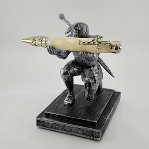 May include: A decorative pen held by a kneeling knight statue. The pen has a cream-colored barrel with gold-colored accents and a pointed tip. The knight statue is silver-colored and rests on a rectangular base.