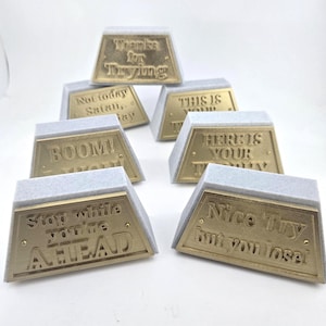 May include: A collection of gold-colored, trapezoid-shaped desk accessories with various engraved phrases like "Thanks for Trying" and "Nice Try but you lose!" Each piece has a light gray base.
