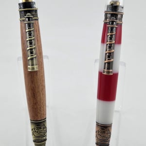 May include: Two decorative lighthouse-themed pens. One pen features a wooden barrel with brass accents, while the other has a red and white striped barrel. Both pens have a lighthouse-shaped top and a detailed ship design at the tip.