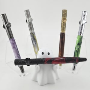 May include: A collection of five decorative pens in various colors and patterns, including purple, yellow, green, and red. The pens are displayed in a white ghost-shaped pen holder. The pens have silver and gold accents.
