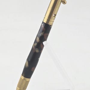 May include: A handcrafted pen with a brass finish and a patterned barrel in shades of brown, beige, and black. The pen features a bolt-action mechanism and a textured grip. It is displayed on a clear acrylic stand.