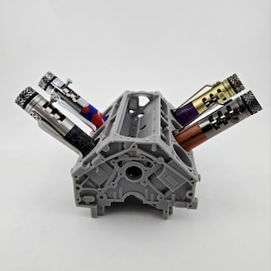 May include: A gray, 3D-printed engine block pen holder with four pens. The pens have various designs, including black, silver, red, purple, and gold accents. The pen holder is a unique desk accessory.