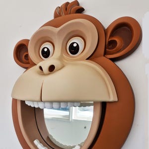 Puede incluir: Un espejo de pared con forma de cara de mono marrón con una gran sonrisa de dientes. El espejo es redondo y tiene un marco blanco. Los ojos del mono son grandes y negros, y su nariz es marrón.