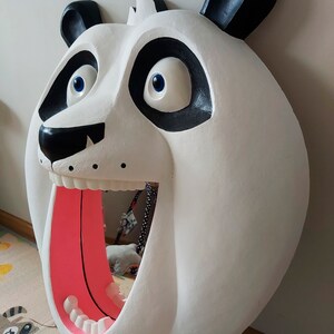 Puede incluir: Una gran escultura de cabeza de panda blanca con detalles negros alrededor de los ojos, las orejas y la nariz. La boca del panda est&aacute; abierta, revelando una lengua rosa y dientes blancos. Los ojos son azules. La escultura est&aacute; montada en una pared.