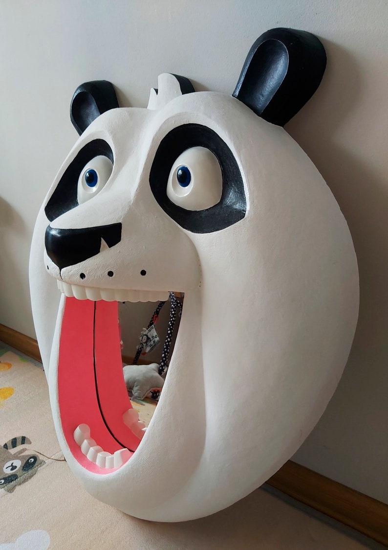 Puede incluir: Una gran escultura de cabeza de panda blanca con detalles negros alrededor de los ojos, las orejas y la nariz. La boca del panda est&aacute; abierta, revelando una lengua rosa y dientes blancos. Los ojos son azules. La escultura est&aacute; montada en una pared.