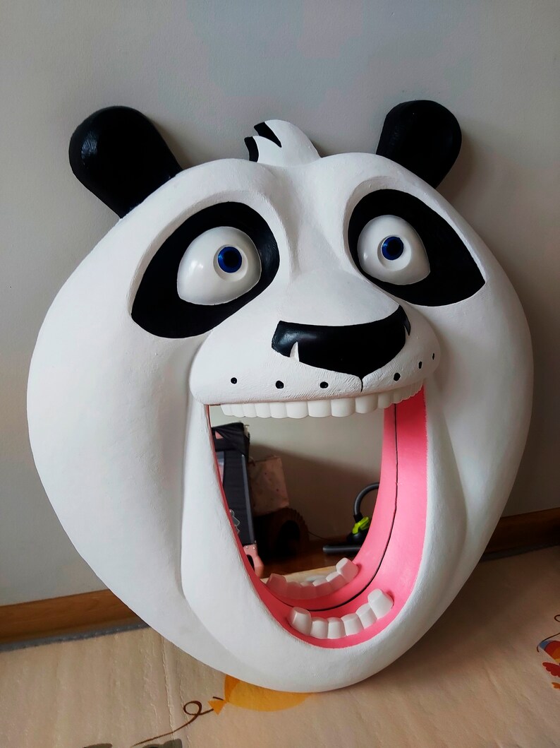 Puede incluir: Una gran cabeza de oso panda blanco con orejas y marcas oculares negras. El panda tiene la boca muy abierta con el interior rosa, revelando dientes blancos. Los ojos son azules. Este art&iacute;culo decorativo es probablemente un colgante de pared o un espejo.