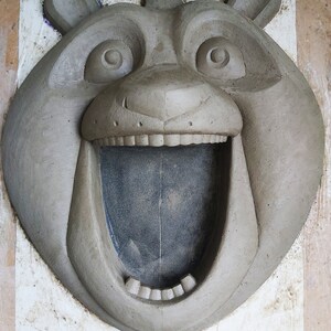 Puede incluir: Una escultura de arcilla gris de la cabeza de un oso con la boca abierta, revelando dientes y un interior oscuro. El oso tiene ojos y orejas grandes y redondos. La escultura est&aacute; sobre una superficie blanca.