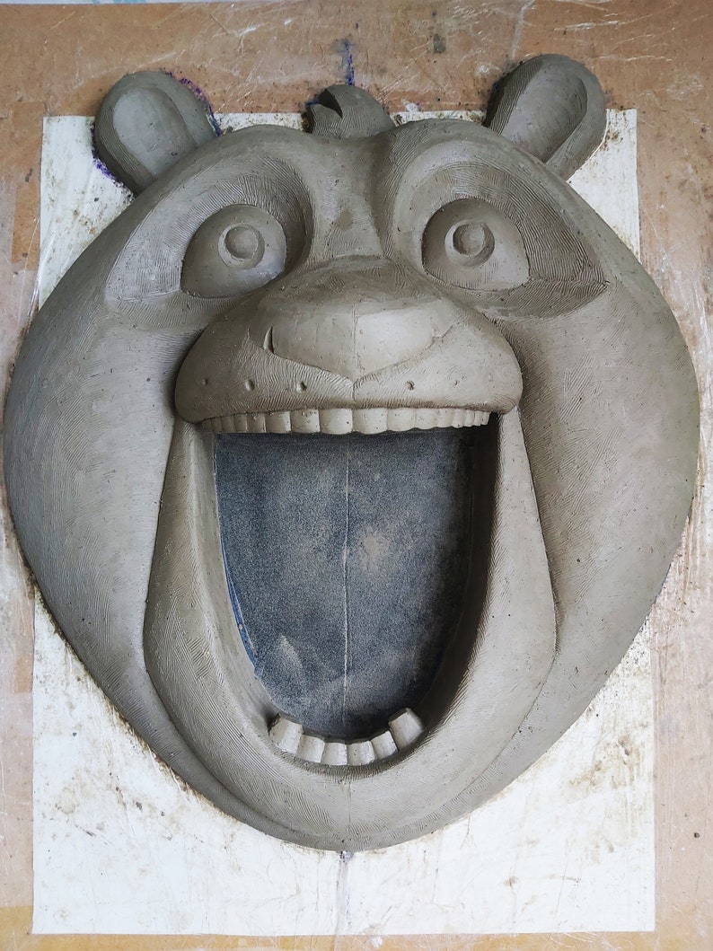 Puede incluir: Una escultura de arcilla gris de la cabeza de un oso con la boca abierta, revelando dientes y un interior oscuro. El oso tiene ojos y orejas grandes y redondos. La escultura est&aacute; sobre una superficie blanca.