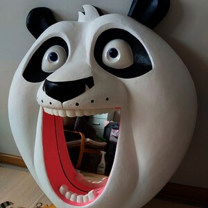 Puede incluir: Un gran espejo blanco con forma de oso panda con detalles negros alrededor de los ojos y las orejas. La boca del panda est&aacute; abierta, revelando un interior rosa y dientes. El espejo refleja la habitaci&oacute;n.