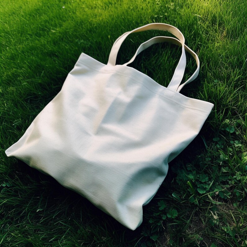 Pu&ograve; includere: Una borsa tote color crema con manici abbinati &egrave; appoggiata su erba verde. La borsa &egrave; realizzata in un tessuto robusto e sembra vuota. I manici sono abbastanza lunghi da essere portati a spalla.