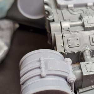 May include: Close-up of a grey plastic model kit component. The cylindrical piece has ribbed detailing and is connected to a larger, angular structure. The model appears to be part of a larger vehicle or structure.