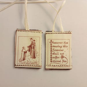 May include: Two white fabric scapulars with brown stitching and text. The left scapular features an image of Our Lady of Mount Carmel and Saint Simon Stock. The right scapular features the text "Whosoever dies wearing this Scapular, shall not suffer eternal fire".