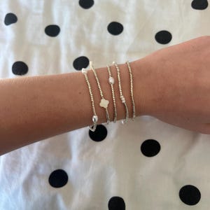 May include: Several silver beaded bracelets, each featuring pearls and a white clover-shaped charm. The bracelets are presented on a white fabric with black polka dots, highlighting their intricate details.