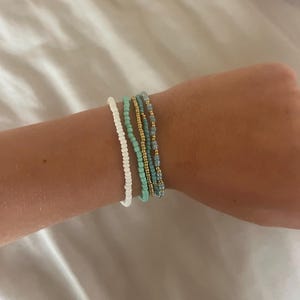 May include: A selection of beaded bracelets in shades of white, turquoise, gold, and blue. The bracelets are worn together on a wrist, displaying the different colours and textures of the beads. The beads are measured in millimetres.