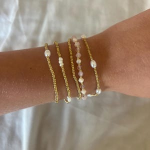 May include: Several gold beaded bracelets, each with different decorative elements. Some have white pearls, others small white beads, and one features pink crystal beads. The bracelets are shown on a wrist against a white backdrop.