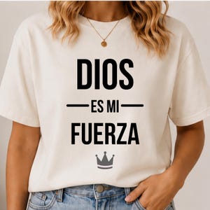 Puede incluir: Camiseta color crema con el texto en español "DIOS ES MI FUERZA" en letras negras en negrita. Un pequeño gráfico de corona gris está debajo del texto. La camiseta tiene mangas cortas y cuello redondo.