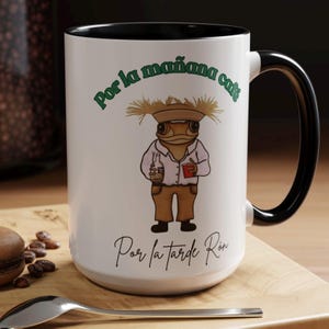 May include: White ceramic mug with a black interior and handle. The mug features a cartoon frog character wearing a hat and holding a bottle and a cup. Text in green and black reads "Por la mañana café" and "Por la tarde Ron".