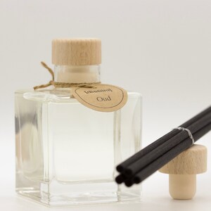 Op de afbeelding: Een heldere glazen diffuserfles met een houten dop en een label met de tekst "Oud". Een bundel zwarte diffuserstokjes en een houten stopper liggen naast de fles. De diffuser is waarschijnlijk voor huisgeur.