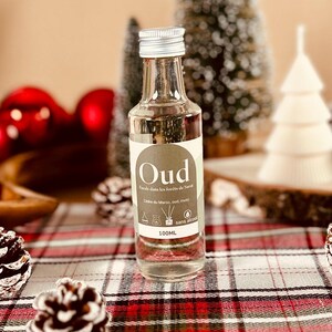 以下が含まれることがあります： フランス語で「Oud」と書かれたラベルが付いた、銀色のキャップが付いた透明なガラス瓶。瓶には透明な液体が入っており、松ぼっくりやミニチュアの木など、クリスマスの装飾を背景に配置されています。100mlの液体が入っています。