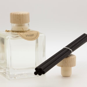 Op de afbeelding: Een heldere glazen diffuserfles met een houten dop en een label met de tekst "Hygge". Een bundel zwarte diffuserstokjes rust op een houten stopper. De diffuser is ontworpen voor huisgeur.