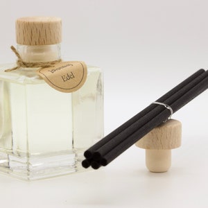 Può includere: Un flacone diffusore in vetro trasparente con tappo in legno e una piccola etichetta marrone con la scritta "Jonah's Edel". Un fascio di bastoncini diffusori neri poggia su un tappo di legno. La bottiglia è riempita di liquido trasparente.