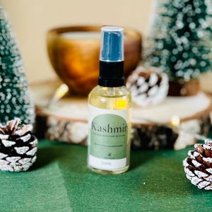 Op de afbeelding: Een heldere glazen fles met een zwarte en blauwe sproeikop, met het label "Kashmir". De fles is gevuld met een lichtgele vloeistof. De fles is omgeven door dennenappels, kleine kerstbomen en een houten kom op een groene ondergrond.