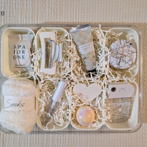 May include: A white spa gift set in a clear tray, filled with various self-care items. Includes a tube of hand cream, a bath bomb, a heart-shaped item, a pair of socks, and other beauty products. The text "SPA FOR ONE" and "Socks" are visible.