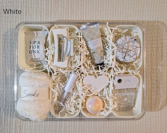 May include: A white spa gift set in a clear tray, filled with various self-care items. Includes a tube of hand cream, a bath bomb, a heart-shaped item, a pair of socks, and other beauty products. The text "SPA FOR ONE" and "Socks" are visible.
