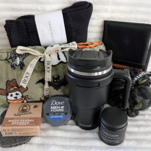 May include: A gift set containing a pair of black socks, a black notebook, a green fleece blanket with Halloween patterns tied with a ribbon, a black travel mug, a bar of Wood Barrel Bourbon soap, a Dove cream tin, a small black candle, and a bath sponge.