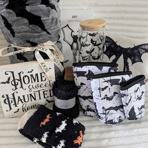 May include: A collection of items featuring bat motifs including a gray plush blanket, a glass tumbler with a bamboo lid, a white wooden sign reading Home sweet HAUNTED home, a beige zippered pouch, three white and black sleeves, and fuzzy socks.