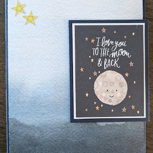 May include: A handmade greeting card with a watercolor background in shades of blue and gray. The card features a smaller rectangular insert with a dark blue background, a smiling moon, stars, and the text "I love you to the moon & back."