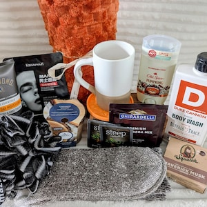 May include: A gift set containing a white and tan ceramic mug, a black and white mesh sponge, grey socks, a Gold Bond lotion tube, a Kissmwah face mask, a candle, Old Spice deodorant, Duke Cannon body wash, Ghirardelli cocoa, and Dr. Squatch soap.