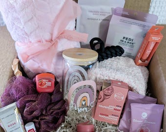 May include: A gift box with a pink theme, filled with self-care items. Includes a pink towel, lip balms, hand cream, a glass jar, a loofah, a soap, and other beauty products. The box is filled with paper filler.
