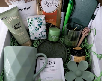 May include: A gift box filled with self-care items. Includes a green mug, fuzzy socks, a tube of Gold Bond Cedar Rain lotion, a Ferrero Rocher chocolate box, a notebook, pens, a candle, and a flower-shaped soap.