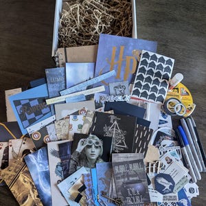 May include: An open cardboard box filled with Harry Potter-themed stationery, stickers, and ephemera. Includes cards, notebooks, pens, and decorative tape. The items are arranged on a wooden surface, with a variety of colors and designs.