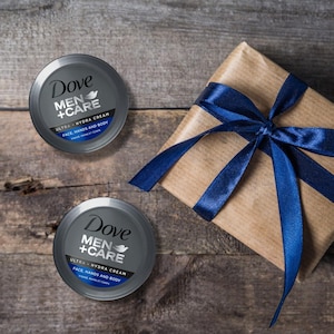 May include: Two Dove Men+Care Ultra Hydra Cream containers with silver lids and blue accents, alongside a wrapped gift with a blue ribbon. The cream is for face, hands, and body. The items are arranged on a rustic wooden surface.