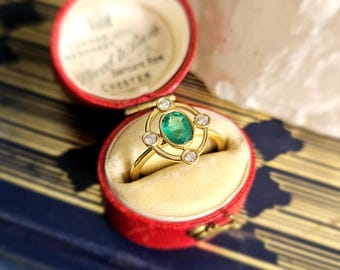 May include: A gold ring with a large emerald green gemstone surrounded by smaller diamonds. The ring is in a red velvet ring box.