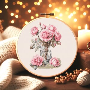 May include: A cross-stitch embroidery hoop with a floral design. The design features a bouquet of pink roses in a clear glass vase. The roses are surrounded by green leaves and stems.