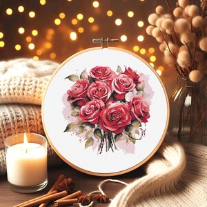 May include: A round embroidery hoop with a white background and a bouquet of red roses with green leaves. The roses are arranged in a circular pattern and have a watercolor effect.
