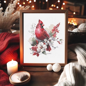 Puede incluir: Un bordado de punto de cruz enmarcado de un cardenal rojo posado entre flores blancas y rojas. La obra de arte está en un marco de madera, con una estética cálida y acogedora. La imagen está sobre una superficie de madera con una vela encendida y ovillos de lana.
