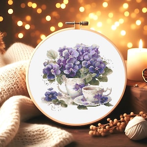 May include: A cross-stitch embroidery hoop with a white background featuring a purple flower arrangement in a white teacup with a saucer and a small teacup with a saucer. The teacups are filled with a brown liquid.