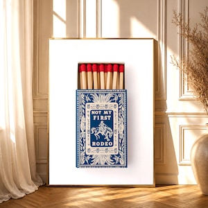 Puede incluir: Obra de arte enmarcada que muestra una caja de cerillas azul con el texto "NOT MY FIRST RODEO" y un vaquero a caballo. La caja de cerillas contiene cerillas con punta roja. La obra está sobre un fondo blanco con un marco dorado.