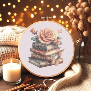May include: A cross-stitch embroidery hoop featuring a stack of books topped with a large rose and smaller buds. Butterflies are also included in the design. The color palette includes shades of pink, beige, and blue.