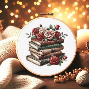 May include: Cross-stitch embroidery in a wooden hoop features a stack of books adorned with red and white roses and green leaves. The books are in shades of brown and green. The background is blurred with warm, golden bokeh lights.