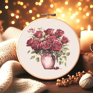 May include: A cross-stitch embroidery hoop with a design of a bouquet of red roses in a brown vase. The roses are arranged in a cluster with green leaves and stems.